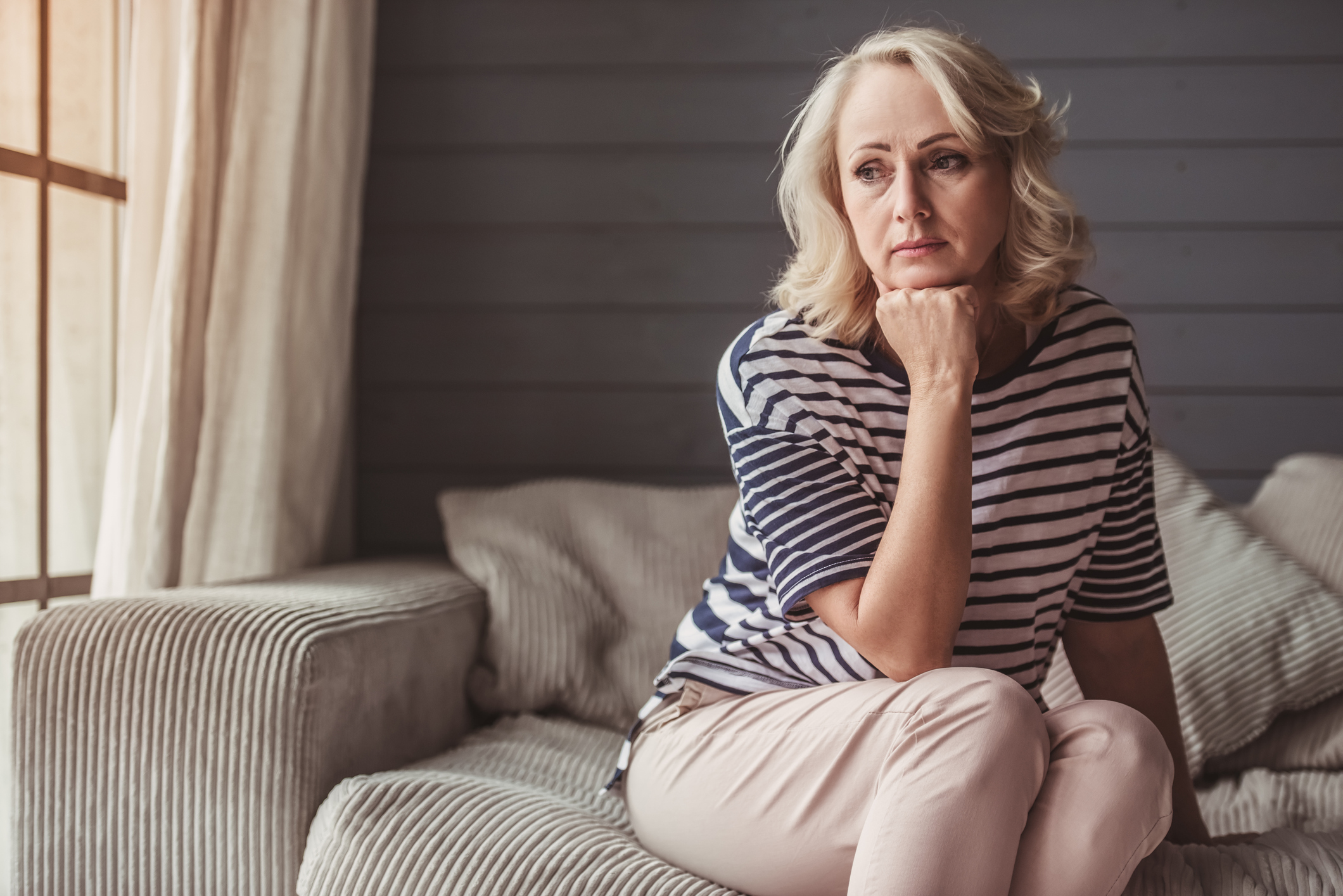 Beautiful sad senior woman is leaning on her hand and looking downward while sitting on couch at home