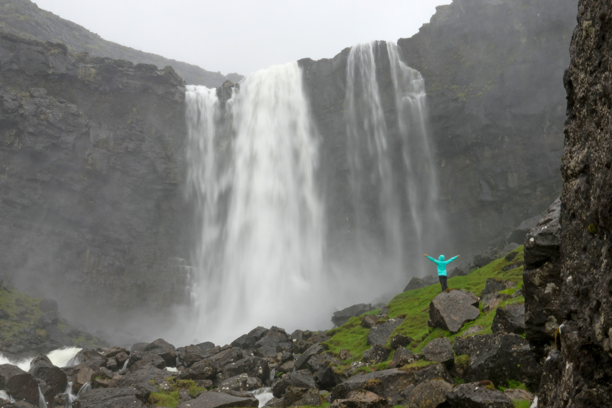 Happy young female tourist with her arms raised looks at the cool waterfall