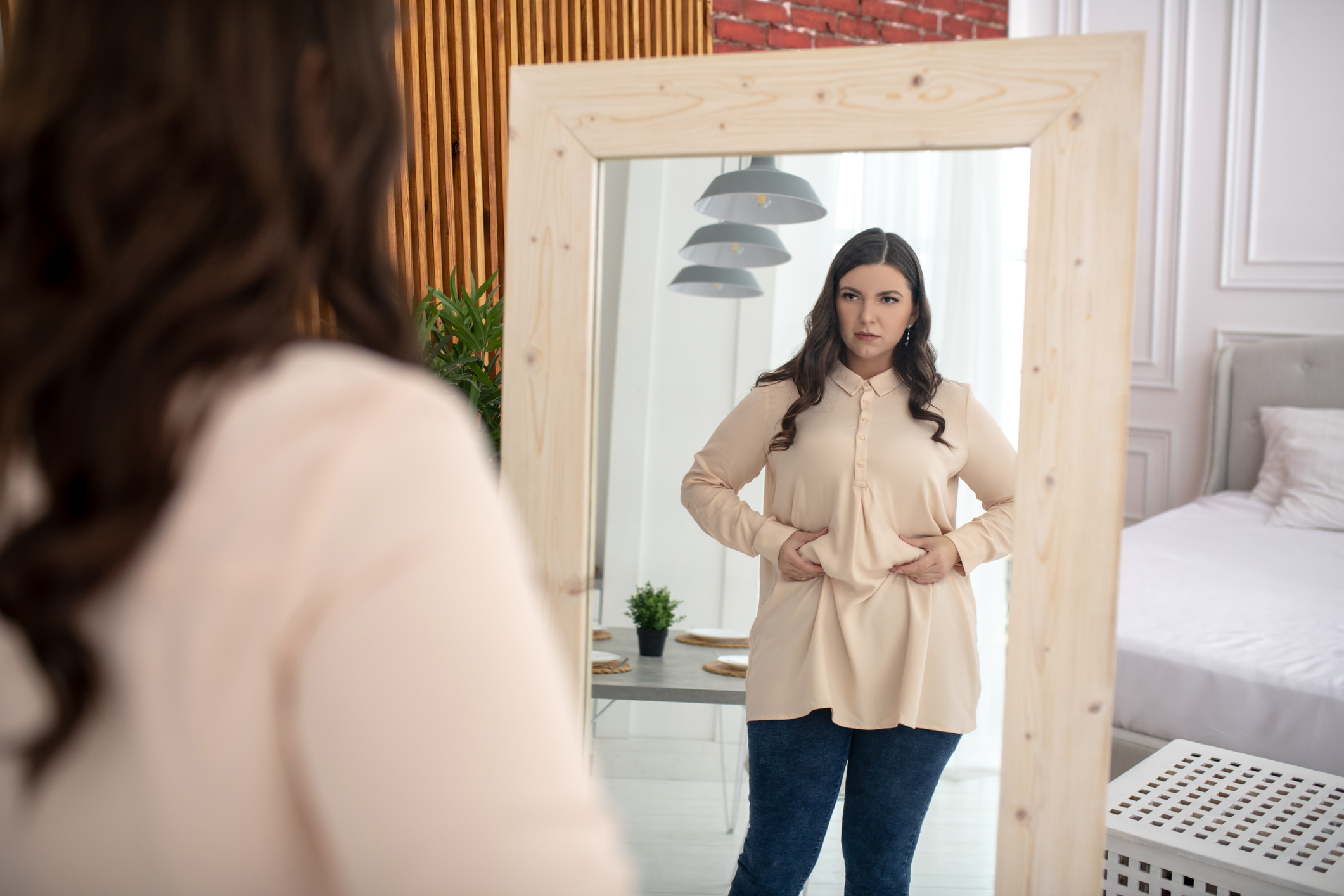 Young woman in a beige blouse squeezing fat on her belly while looking in a mirror